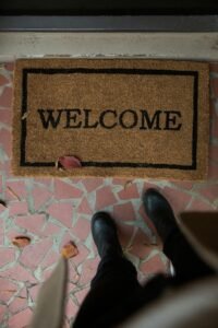 Top-down perspective of a welcome mat and black boots on a tiled floor with fallen leaves.